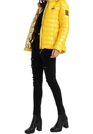 Person iført gul polstret jakke med guldforing, sorte revnede skinny jeans og sorte ankelstøvler med hæl, stående mod hvid baggrund.