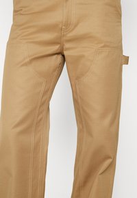 Pantalon de travail couleur sable avec coutures renforcées au niveau des cuisses et une petite boucle sur le côté droit près de la zone de la poche.