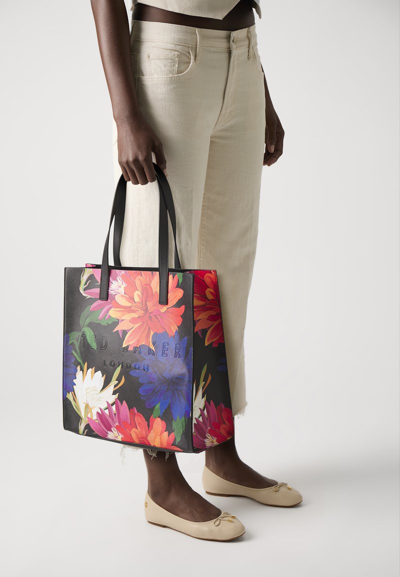 Svart totebag med et levende blomstermønster i rosa, blått og hvitt. Har solide svarte håndtak og preget logo på fronten.