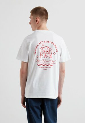 Młody mężczyzna zwrócony tyłem, ubrany w białą koszulkę z czerwoną ilustracją psa i tekstem dotyczącym spodni typu slacks i odzieży rekreacyjnej na plecach.