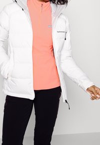 Vrouw in een witte pufferjack over een koraal kleurige rits top en zwarte broek, poseren met één hand in de jaszak en de andere uitgestrekt.