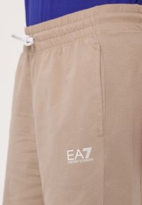 Beige treningsshorts med elastisk midjebånd og snøring. Har side-lommer og en hvit EA7 Emporio Armani-logo. Myk stofftekstur.