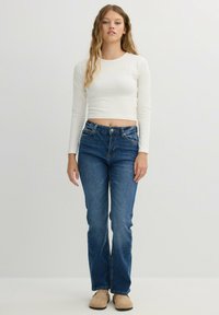 Top corto de manga larga blanca combinado con jeans ajustados azules y zapatos beige. Diseño de pierna recta con textura de denim clásico y un ligero desgaste.