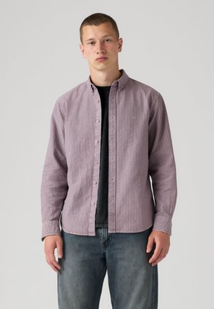 Jeune homme portant une chemise boutonnée rayée violette sur un t-shirt noir et un jean bleu, debout devant un fond clair uni.