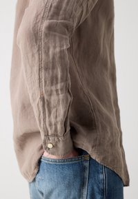 Personne portant une chemise beige froissée à manches longues avec des poignets boutonnés et un jean bleu, une main dans la poche.