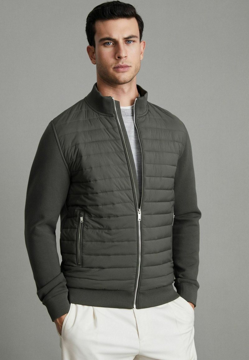 Veste bomber pour homme en vert foncé, avec un devant matelassé, des manches lisses, un col haut et des poches zippées pour plus de praticité.