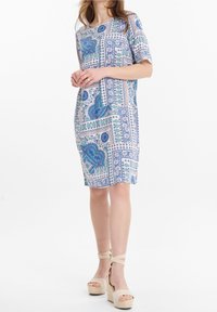 Frau in einem knielangen, kurzärmeligen Kleid mit blauen und grünen Paisleymustern, kombiniert mit beigen Plateausandalen mit Knöchelriemen.