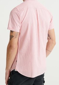 Camicia a maniche corte di colore rosa chiaro con righe verticali, tessuto testurizzato, colletto button-down e orlo curvo con spacco laterale.