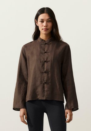 Kobieta o długich ciemnych włosach nosi brązową kurtkę z kołnierzem mandarynki i zapięciami w stylu „żabek”, stoi na tle jednolitego jasnego tła.