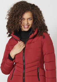 Sarkans pūkains jaka ar rāvējslēdzi priekšpusē, pārklāta ar kviltētām tekstūrām, melniem akcentiem un augstu apkakli. Iekļauj sānu kabatas un elpojošas sānu plāksnes.