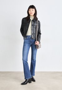 Giacca in pelle nera con chiusura a bottoni e due tasche frontali, abbinata a un dolcevita verde chiaro, jeans flare blu e stivali neri con tacco.