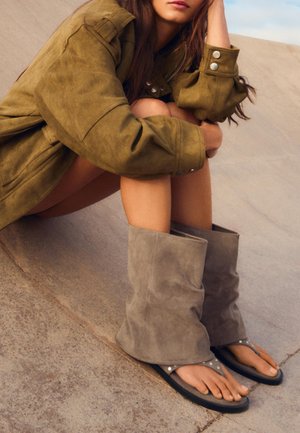 Femeie care poartă o jachetă oversized de culoare măslinie, jambiere gri din piele întoarsă și papuci cu ținte, stând cu genunchii îndoiți pe o pantă de beton.