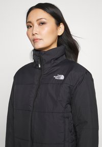 Crna zimska jakna od pufa s visokim ovratnikom, zatvaračem sprijeda i šivenim panelima. Sadrži logo The North Face na lijevoj strani prsiju. Glatka tekstura.