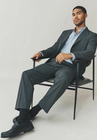 Costume gris avec une finition texturée, chemise rayée blanche et bleue en dessous, mocassins noirs, assis sur une chaise en métal noir contre un fond clair.