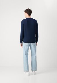 Tengerészkék, hosszú ujjú kötött felső, kerek nyakkal, világoskék, laza farmernadrággal és fehér sportcipővel, hátulról nézve.