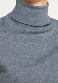 Grå stickad tröja med mönster av strukturerad vävning. Har en hög, ribbad krage och en jämn yta genom hela tröjan.