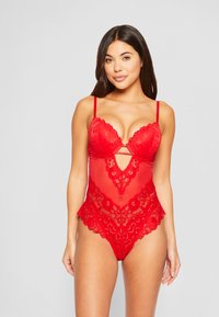 Raudonas nėriniuotas bodis su permatomais tinkliniais intarpais, turintis gilią iškirptę, banguotais kraštais, gėlių raštais ir reguliuojamomis petnešomis.