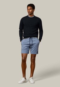 Uomo che indossa un maglione nero a maniche lunghe, camicia a righe, pantaloncini blu con coulisse e scarpe da ginnastica bianche, in piedi con le mani nelle tasche su sfondo neutro.