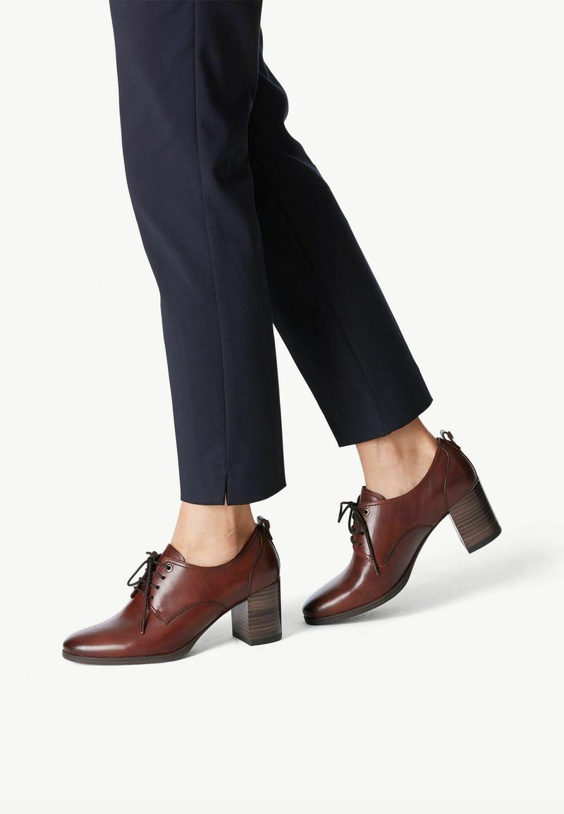 Pantalon bleu marine foncé associé à des chaussures à talons blocs en cuir marron à lacets, visibles de mi-mollet vers le bas, sur fond blanc.