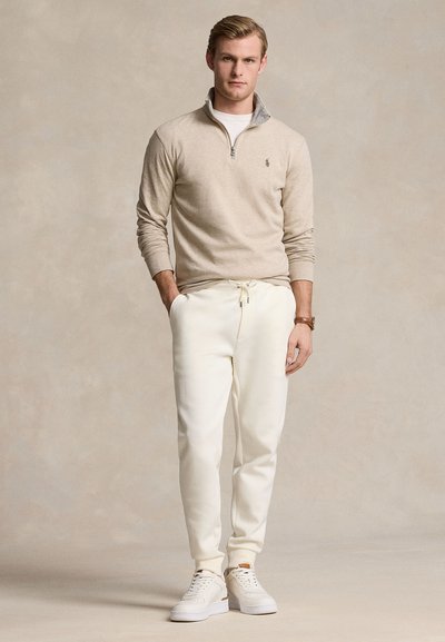 Joven con suéter beige de cremallera parcial, pantalones blancos con cordón, zapatillas blancas y un reloj de pulsera, de pie con las manos en los bolsillos.