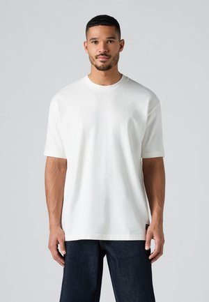 T-shirt bianca a maniche corte con scollo rotondo, realizzata in cotone. Presenta un logo sottile sul petto. Indossata con jeans in denim scuro.