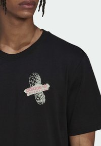 Černé tričko s krátkým rukávem s texturovaným grafickým potiskem podrážky boty a nápisem "adidas ADVENTURE" v růžové barvě.