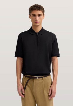 Junger Mann mit schwarzem kurzärmligem Polo mit Reißverschluss und beigen Hosen mit geflochtenem braunem Gürtel, stehend vor einem einfarbigen Hintergrund.