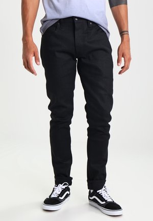 Mann in schwarzer Slim-Fit-Jeans und schwarz-weißen Sneakern, steht mit entspannt an den Seiten hängenden Händen.