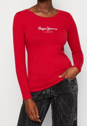 Woman wearing a fitted red long-sleeve shirt with white “Pepe Jeans London” logo and black acid-wash style jeans, hand in pocket.