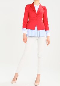 Blazer rouge ajusté avec un revers cranté, deux boutons et des poches latérales, porté sur une chemise bleu clair et associé à un pantalon blanc.