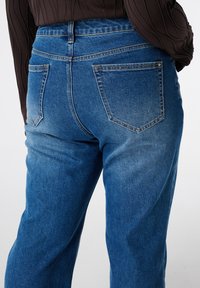 Jean en denim bleu taille haute avec un lavage délavé, cinq poches et des coutures nettes. L'arrière présente deux poches plaquées courbes.