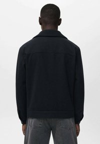 Chaqueta de lana negra con cuello puntiagudo, mangas largas y bolsillos laterales. Textura suave y diseño sencillo, vista desde la parte posterior.