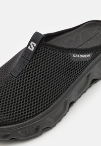 Pantof cu plasă neagră, tip slip-on, cu o suprafață texturată, având un logo gri și o talpă robustă din cauciuc pentru aderență și suport.