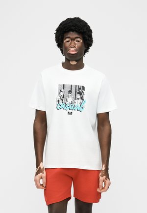 Άνδρας με λεύκη που φοράει λευκό μπλουζάκι με σχέδιο και κόκκινα σορτς, στέκεται μπροστά σε καθαρό φωτεινό φόντο.