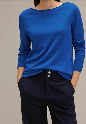 Kobieta ubrana w jasnoniebieski sweter z długim rękawem oraz ciemnogranatowe spodnie z srebrnymi guzikami, stojąca z rękami w kieszeniach.