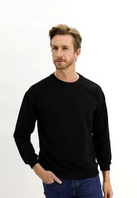 Sudadera negra de manga larga, con cuello redondo, puños acanalados y dobladillo. Llevada con jeans azules, modelo de pie contra un fondo blanco.