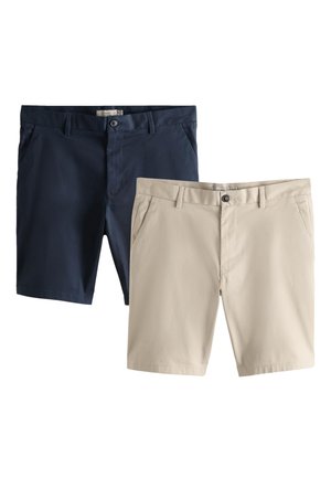 Ung mand iført en sort sweater, beige shorts, hvide sokker og brune sneakers, står med hænderne i lommerne mod en plain væg.