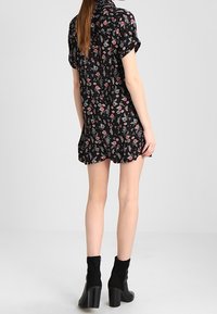 Schwarzes kurzes Kleid mit Blumenmuster, kurzen Ärmeln, gerafftem Saum und einem Kragen. Getragen mit schwarzen Stiefeletten mit einem kräftigen Absatz.