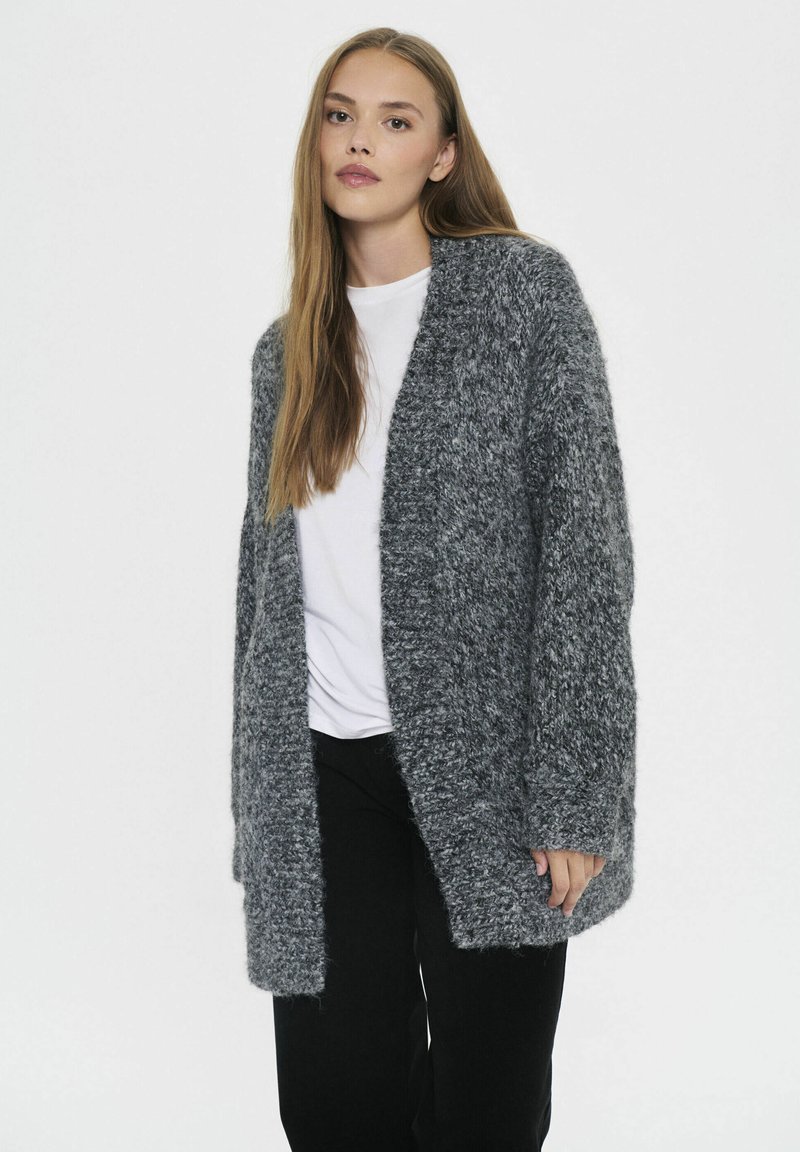Sivý, nadýchaný kardigán vyrobený z textúrovaného materiálu s otvoreným predným dielom. Skombinovaný s bielym topom a čiernymi nohavicami, s dlhými rukávmi.