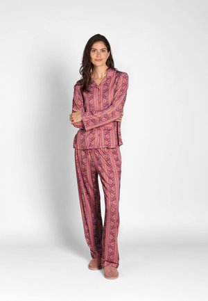 Woman with long dark hair stands with arms crossed, wearing a matching pink patterned long-sleeve top and pants, with pink slippers.