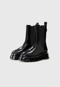 Botas chelsea negras brillantes con paneles laterales elásticos, suela texturizada gruesa y lengüetas en la parte trasera. Material suave con un diseño elegante.