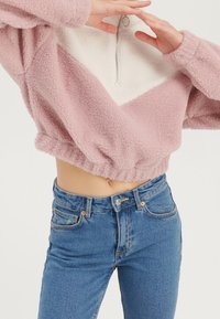 Flauschiger, pinker, kurz geschnittener Pullover mit weißem Chevron-Panel, halbhohem Reißverschlusskragen und elastischem Saum. Kombiniert mit hoch taillierter blauer Denim-Jeans.