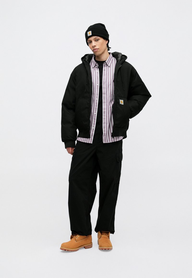 Joven vestido con un gorro negro, chaqueta acolchada negra, camiseta a rayas moradas, pantalones cargo negros y botas de trabajo color beige, de pie contra un fondo blanco.