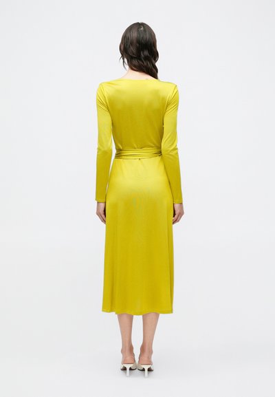 Rochie cu mânecă lungă, lungime până la gleznă, dintr-un material galben aprins cu o textură fină și curea legată în talie, privită din spate.