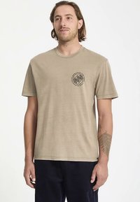 Khakinvärinen lyhythihainen t-paita pehmeästä kankaasta, jossa musta pyöreä graafinen kuvio vasemmassa rinnassa. Pyöreä pääntie, rento istuvuus.