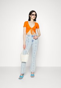 Top cropped laranja com textura canelada e frente amarrada, jeans flare azul-claro, sapatos de salto azuis com tiras, e uma mala branca tipo bucket. Óculos de sol oversized pretos.