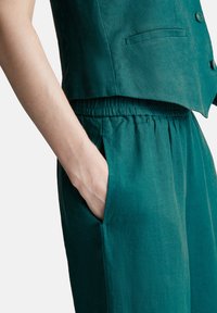 Persona con la mano nella tasca elastica dei pantaloni in lino verde scuro, indossa un gilet abbottonato abbinato.