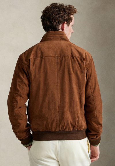 Bărbat cu păr creț, purtând o geacă bomber maro din piele întoarsă și pantaloni crem, stând cu mâinile în buzunare, cu spatele spre privitor.