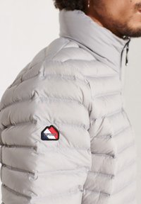 Veste matelassée gris clair avec texture capitonnée, col haut et un écusson logo avec des accents rouge, blanc et bleu sur la manche.