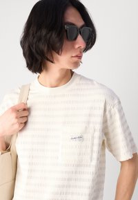 Creme kortærmet t-shirt med teksturerede vandrette striber, der har en lomme med et logo-tag. Modellen bærer en beige taske og har mørke solbriller på.